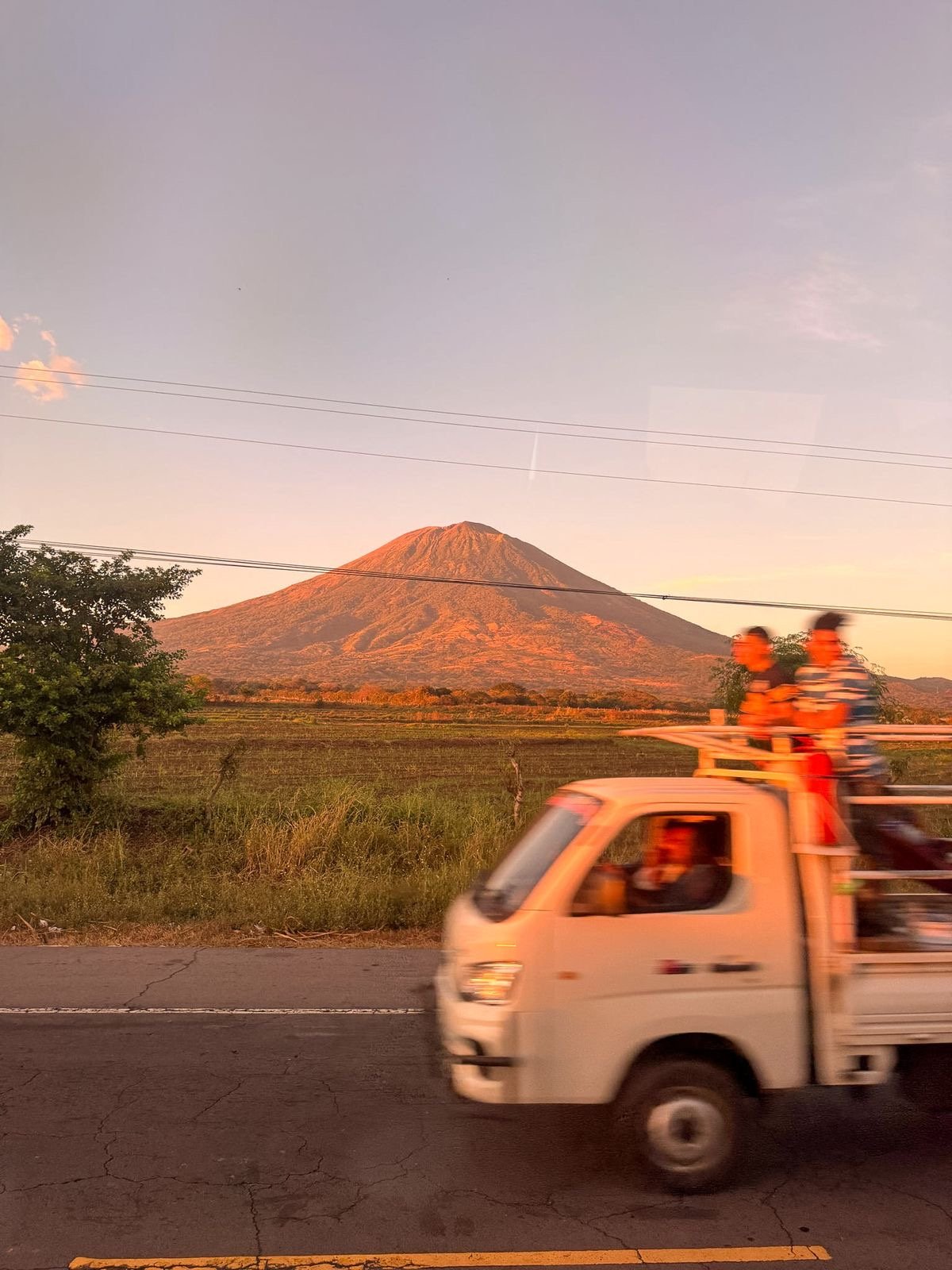 A pickup truck with people riding in the back passes a volcano at sunset in El Salvador